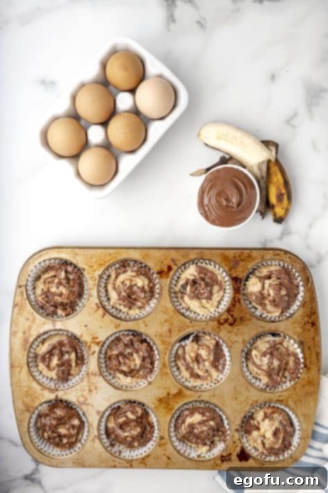 Warmed Nutella being spooned onto the surface of the banana muffin batter in lined muffin cups, awaiting the swirl.
