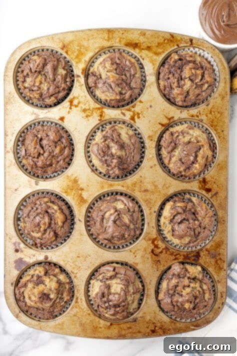 Freshly baked banana Nutella muffins cooling slightly in their muffin tin, exhibiting beautiful golden tops and visible Nutella swirls.