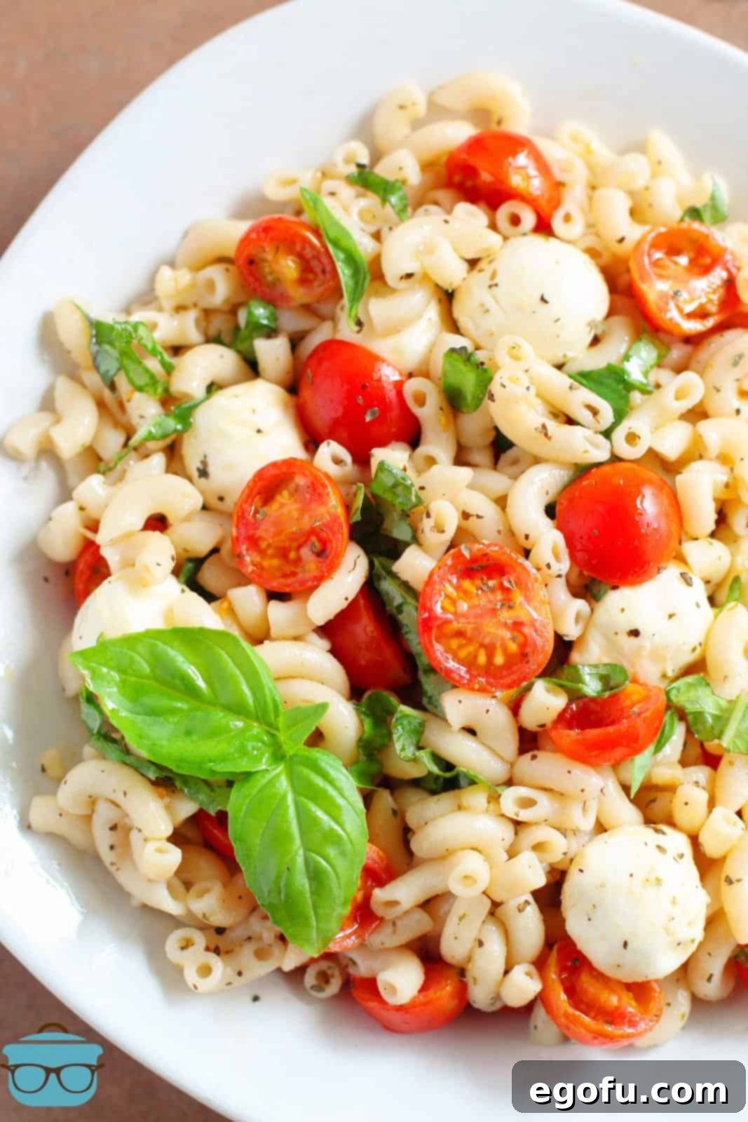 A large white serving dish filled with Caprese Pasta Salad, featuring elbow macaroni, halved cherry tomatoes, small mozzarella balls, and fresh basil leaves, ready to be served.