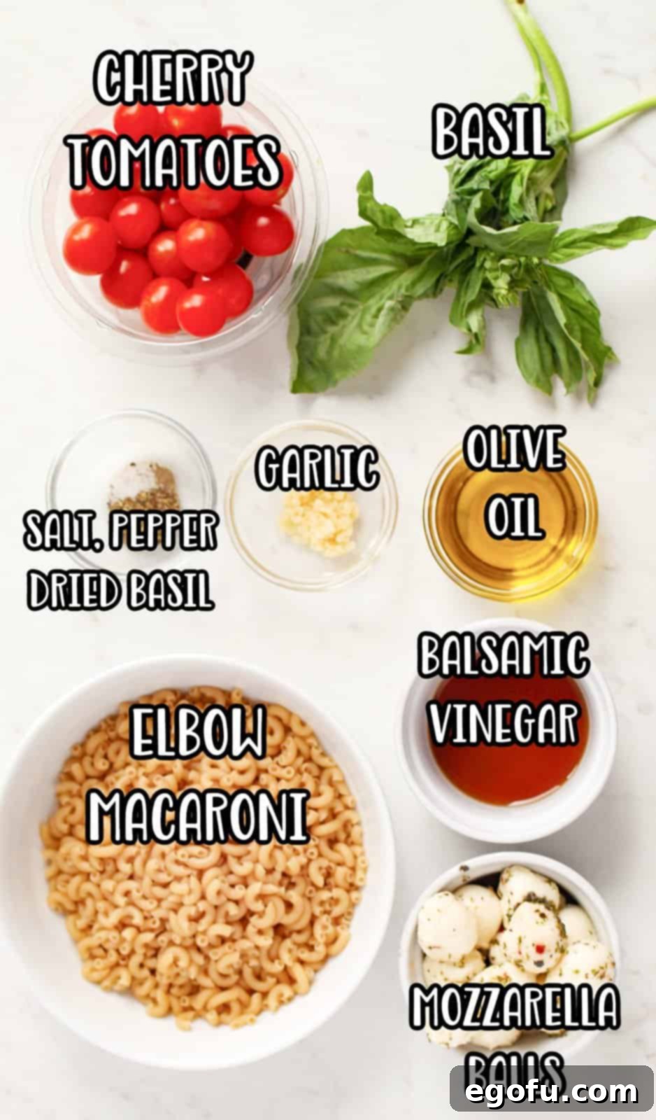 A flat lay of all the Caprese Pasta Salad ingredients: uncooked elbow macaroni, bright red cherry tomatoes, fresh green basil leaves, a bottle of olive oil, a container of marinated mozzarella balls, garlic cloves, a measuring spoon with sugar, a small bowl with black pepper, a small bowl with sea salt, and a bottle of white balsamic vinegar.