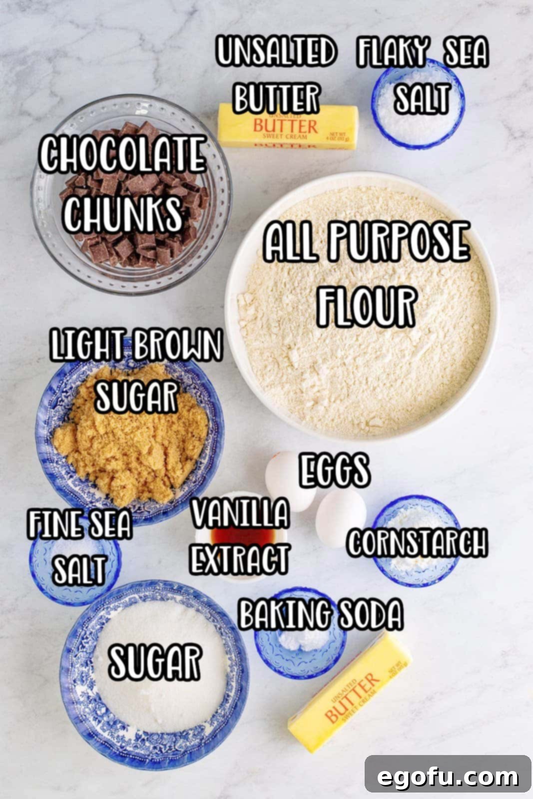 A flat lay photograph displaying all the fresh ingredients for chocolate chunk cookies: a bowl of flour, cornstarch, baking soda, and sea salt, alongside softened butter, granulated sugar, brown sugar, eggs, vanilla extract, a bag of semi-sweet chocolate chunks, and a small dish of flaky sea salt.