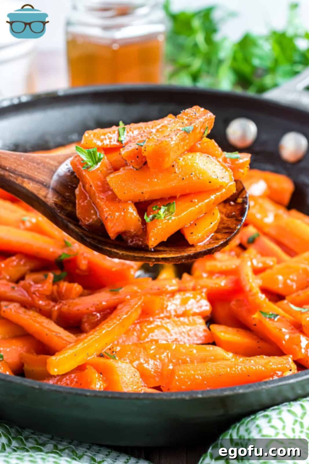 A wooden spoon holding a serving of Honey Glazed Carrots above a skillet full of carrots, showcasing their rich, golden glaze.