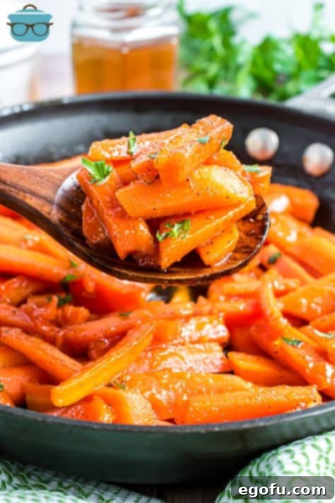 A wooden spoon holding a serving of Honey Glazed Carrots above a skillet full of carrots.