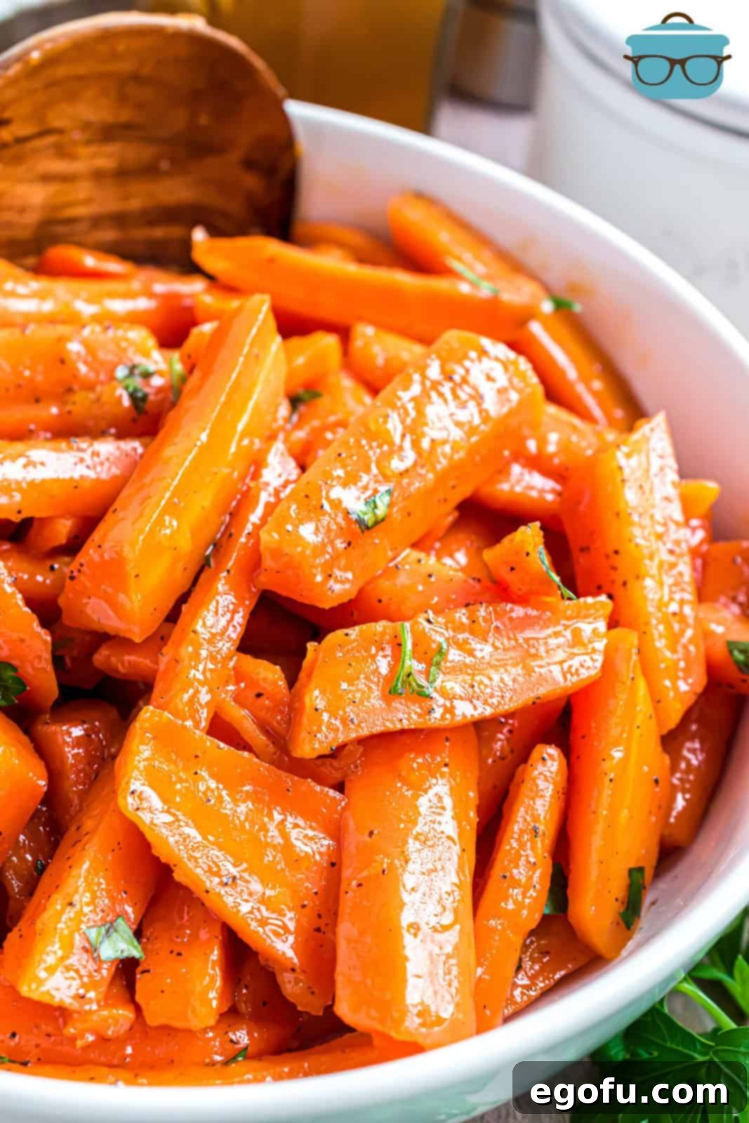 A wooden spoon holding a perfect serving of Honey Glazed Carrots, glistening with their sweet glaze, above a skillet full of more carrots.