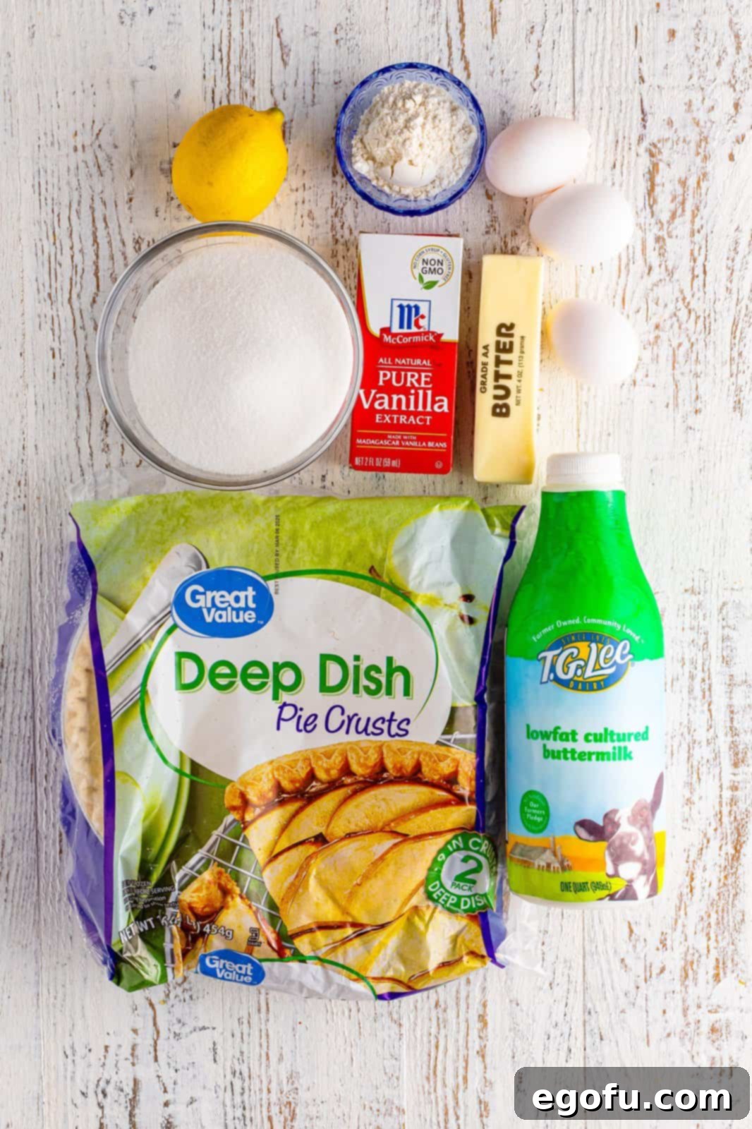 A flat lay photograph showcasing all the ingredients required for Buttermilk Pie: pie crust, granulated sugar, all-purpose flour, large eggs, fresh lemon juice, melted butter, vanilla extract, and buttermilk.