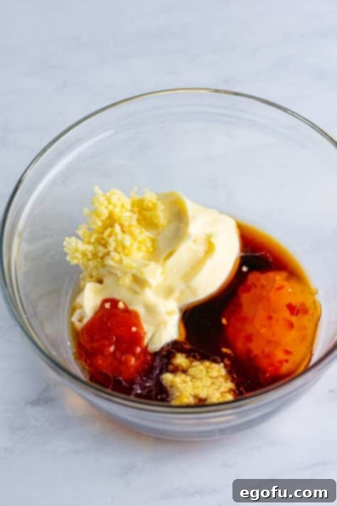 A glass mixing bowl with mayonnaise, rice wine vinegar, sweet chili sauce, honey, soy sauce, garlic, sriracha, ginger, and sesame oil.