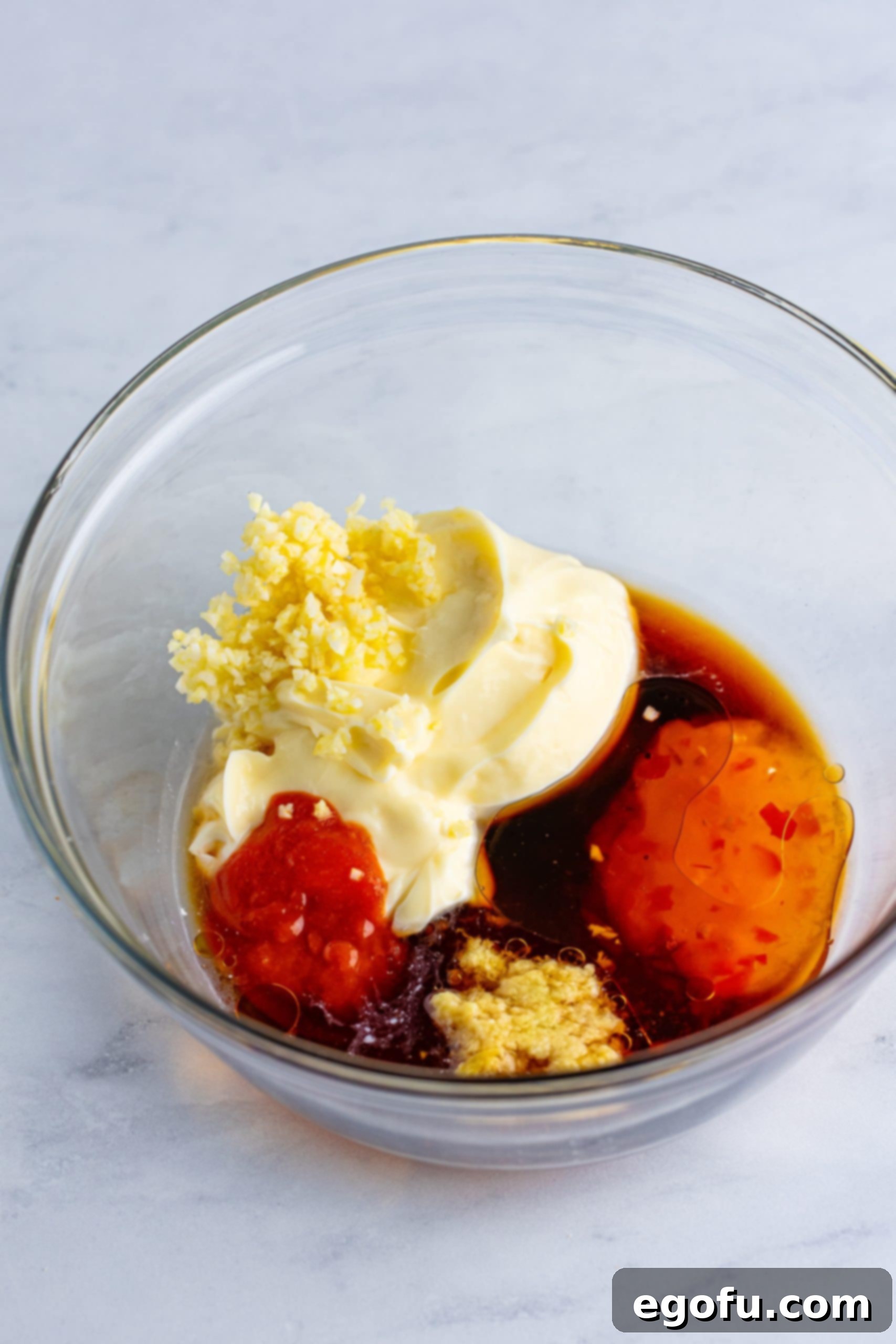 A glass mixing bowl with mayonnaise, rice wine vinegar, sweet chili sauce, honey, soy sauce, garlic, sriracha, ginger, and sesame oil. 