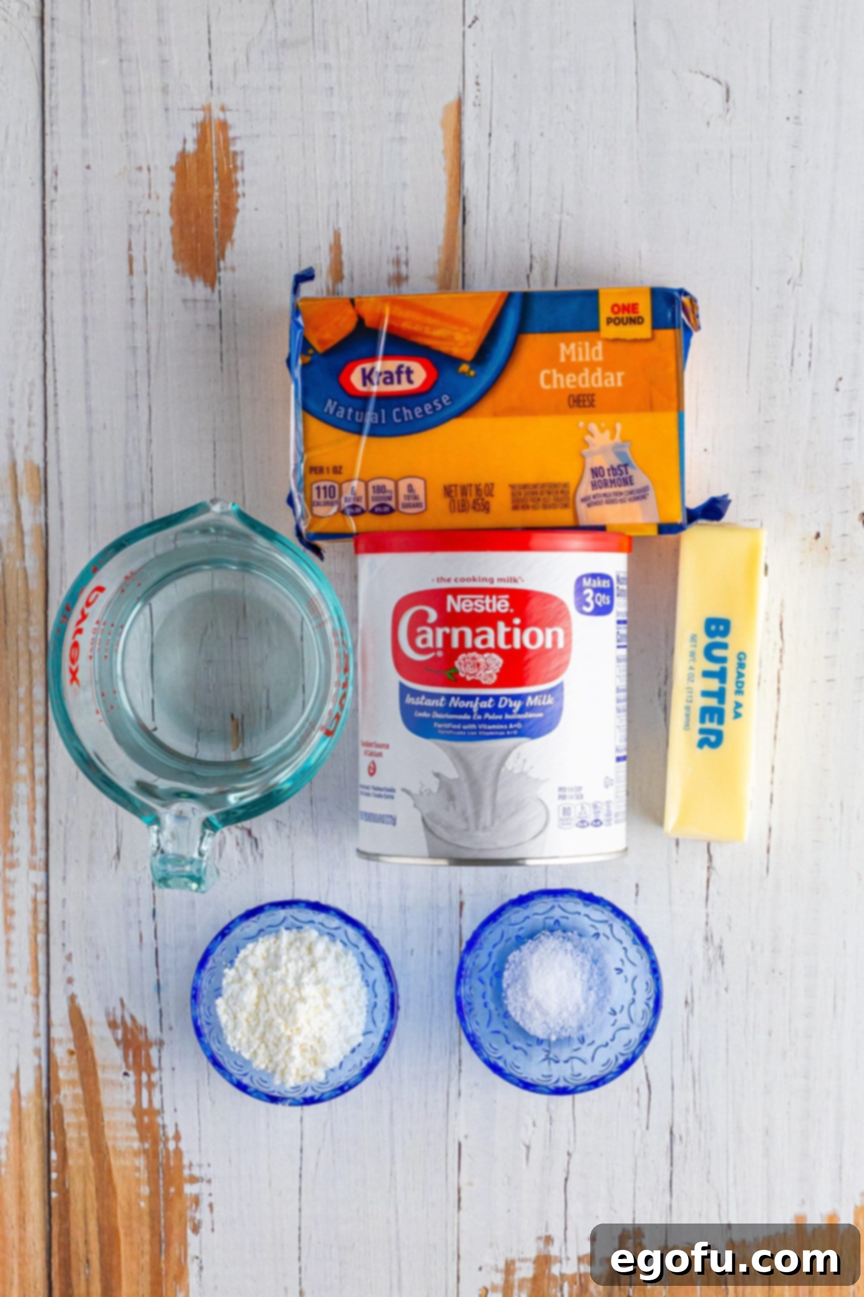 An overhead shot showcasing the six essential ingredients for Homemade Velveeta: a block of mild cheddar cheese, a stick of butter, a small bowl of salt, a measuring cup of water, a container of nonfat milk powder, and a small bowl of cornstarch.