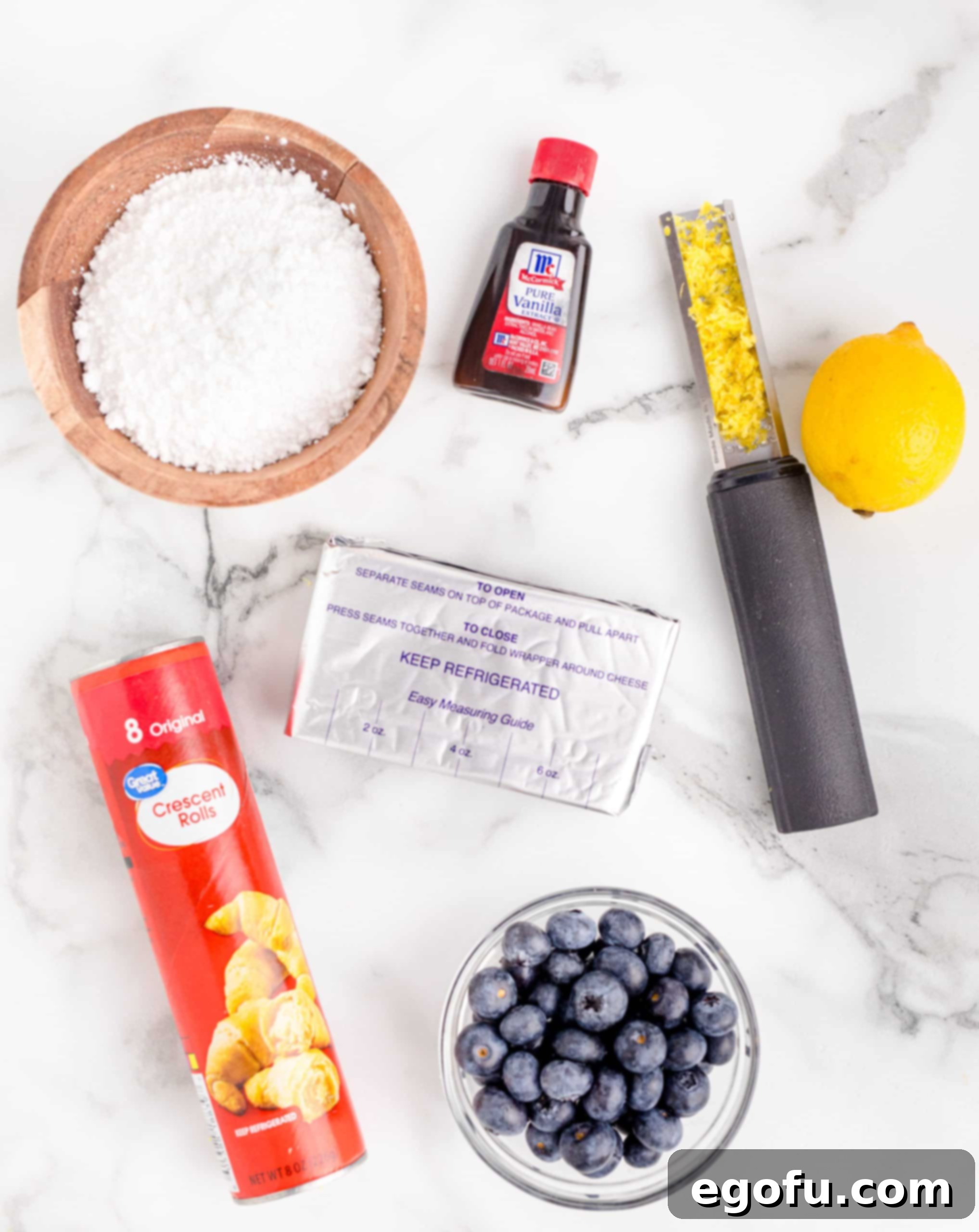 A vibrant flat lay of key ingredients: cream cheese, fresh blueberries, crescent rolls, lemons, vanilla extract, powdered sugar, arranged neatly.