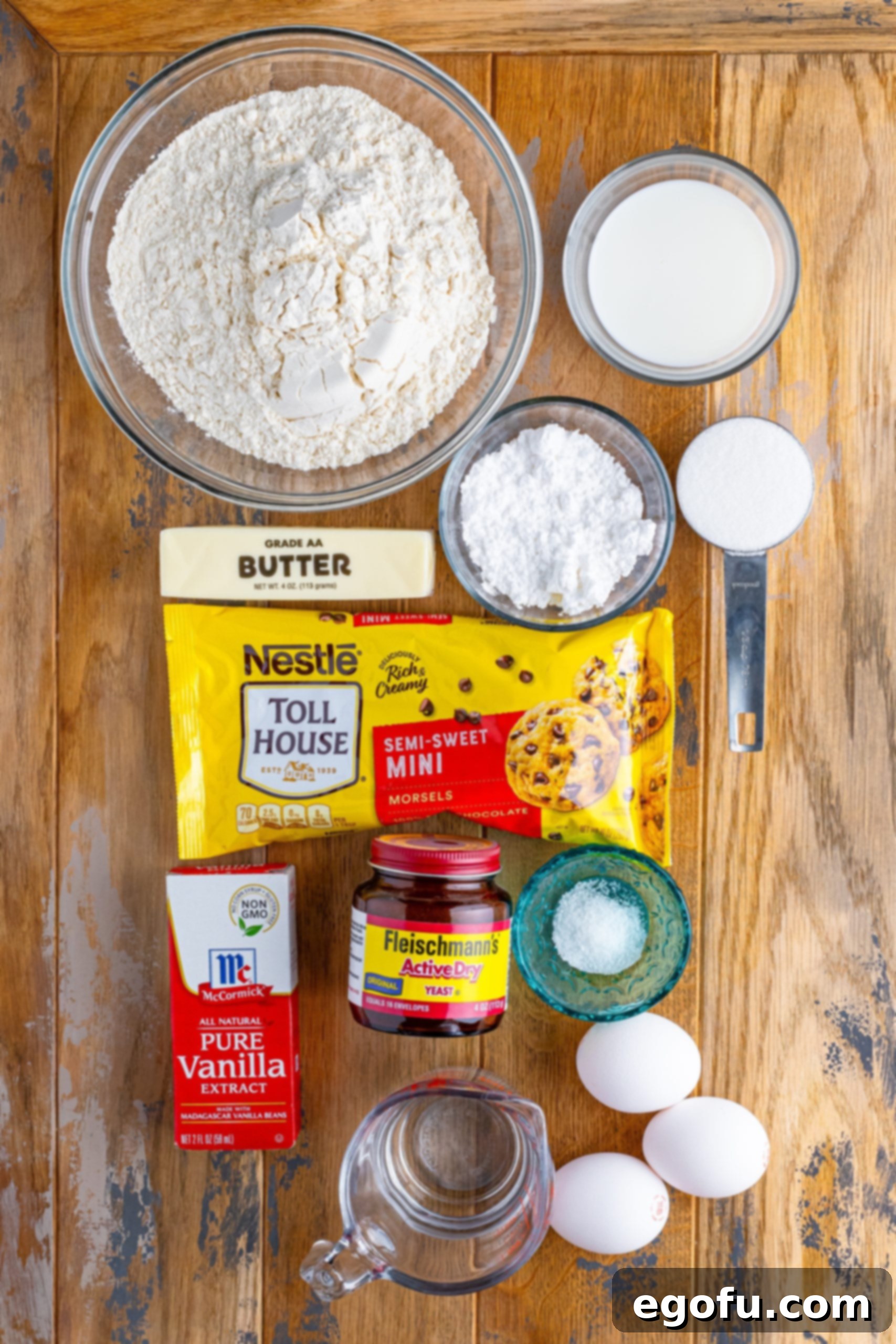 Indulgent Chocolate Hot Cross Buns 4 A flat lay of all the ingredients needed for Hot Cross Buns, including flour, water, yeast, sugar, salt, egg yolks, butter, milk, vanilla extract, mini chocolate chips, and powdered sugar.