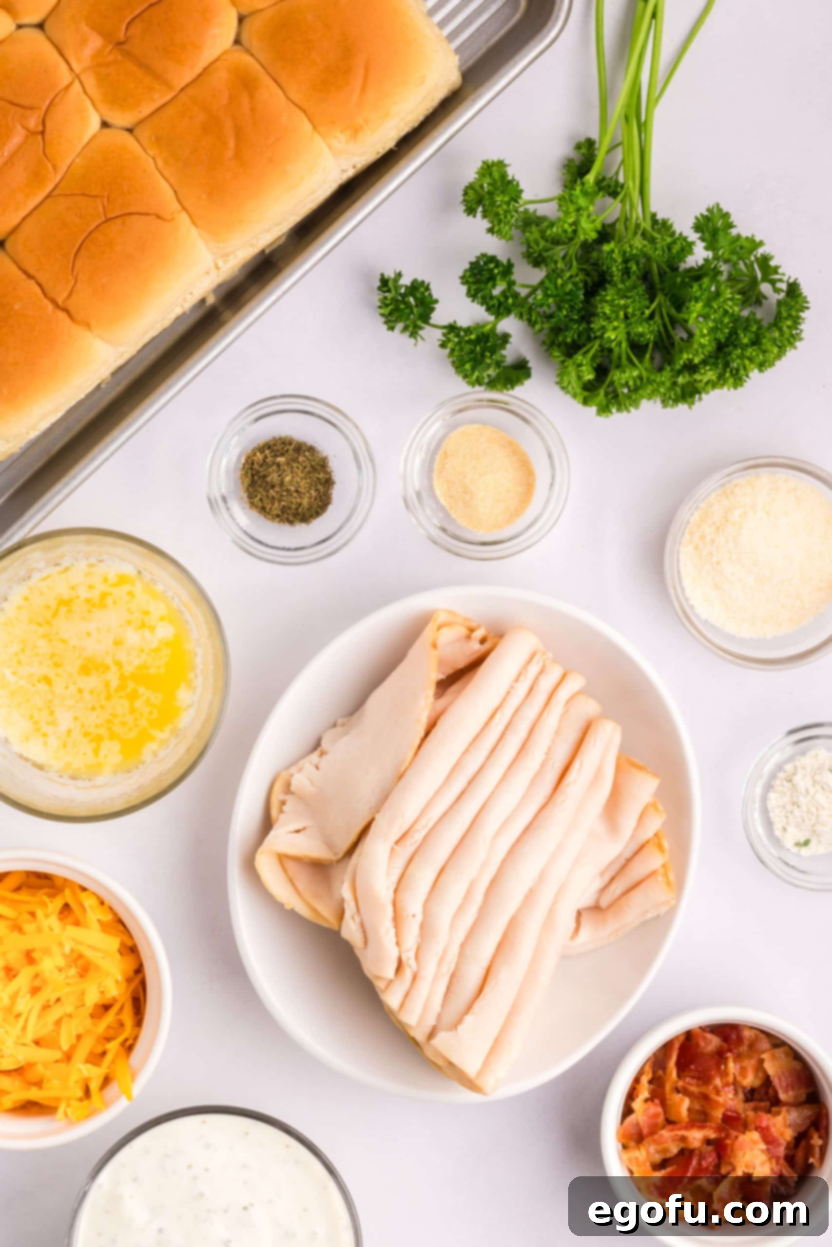 An array of ingredients for Chicken Bacon Ranch Sliders including rolls, butter, parsley, parmesan cheese, ranch seasoning, garlic powder, dill, ranch dressing, deli chicken, bacon, and shredded cheddar cheese.