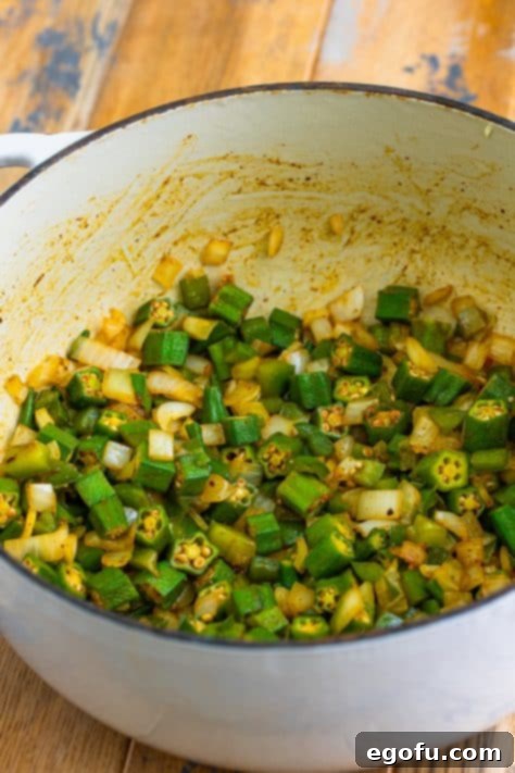 A Dutch oven containing sautéing onion, bell pepper, okra, salt, pepper, and paprika in bacon grease.