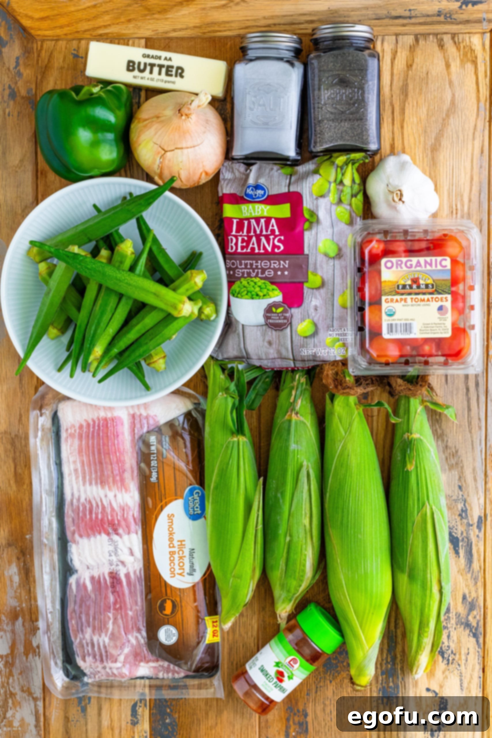 An organized display of fresh ingredients for succotash: corn, lima beans, onion, green bell pepper, butter, salt, okra, paprika, garlic, and grape tomatoes, all neatly arranged and ready for preparation.