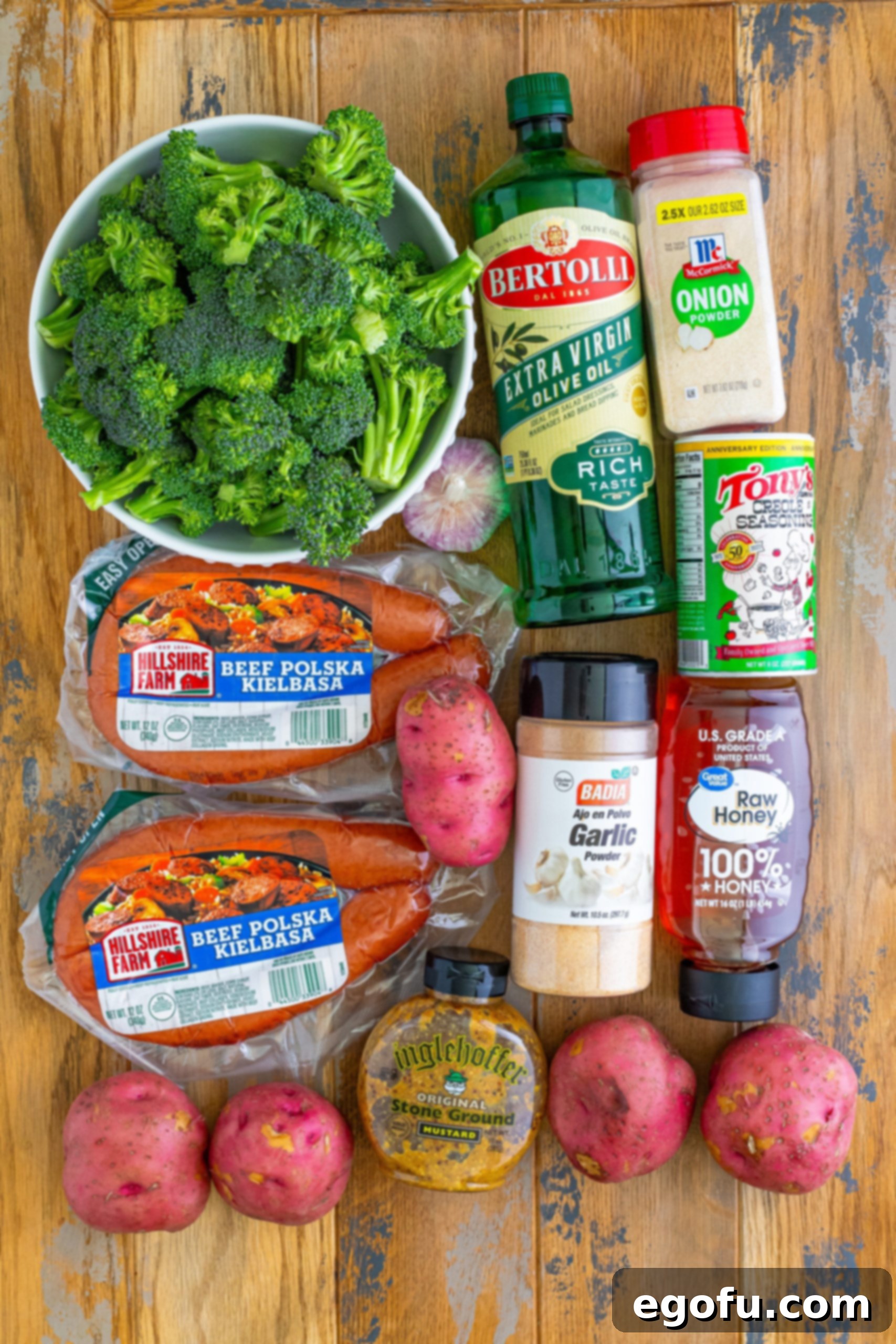 Beef Kielbasa, broccolini, olive oil, red potatoes, creole seasoning, garlic powder, onion powder, honey, stone ground mustard, and garlic cloves arranged for the recipe.