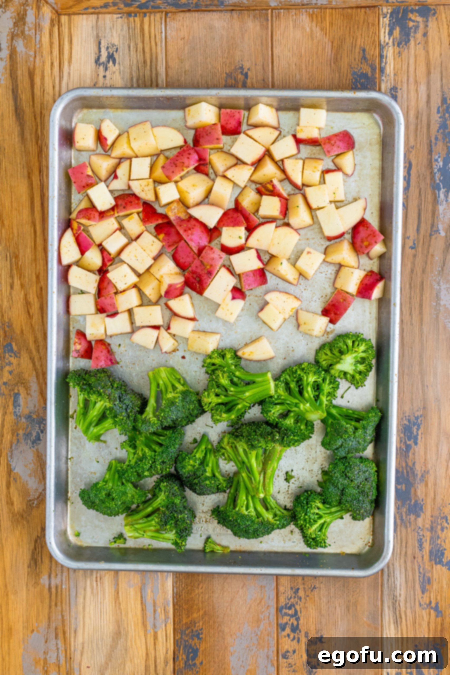 A sheet pan with broccolini and diced potatoes, seasoned and ready for roasting.