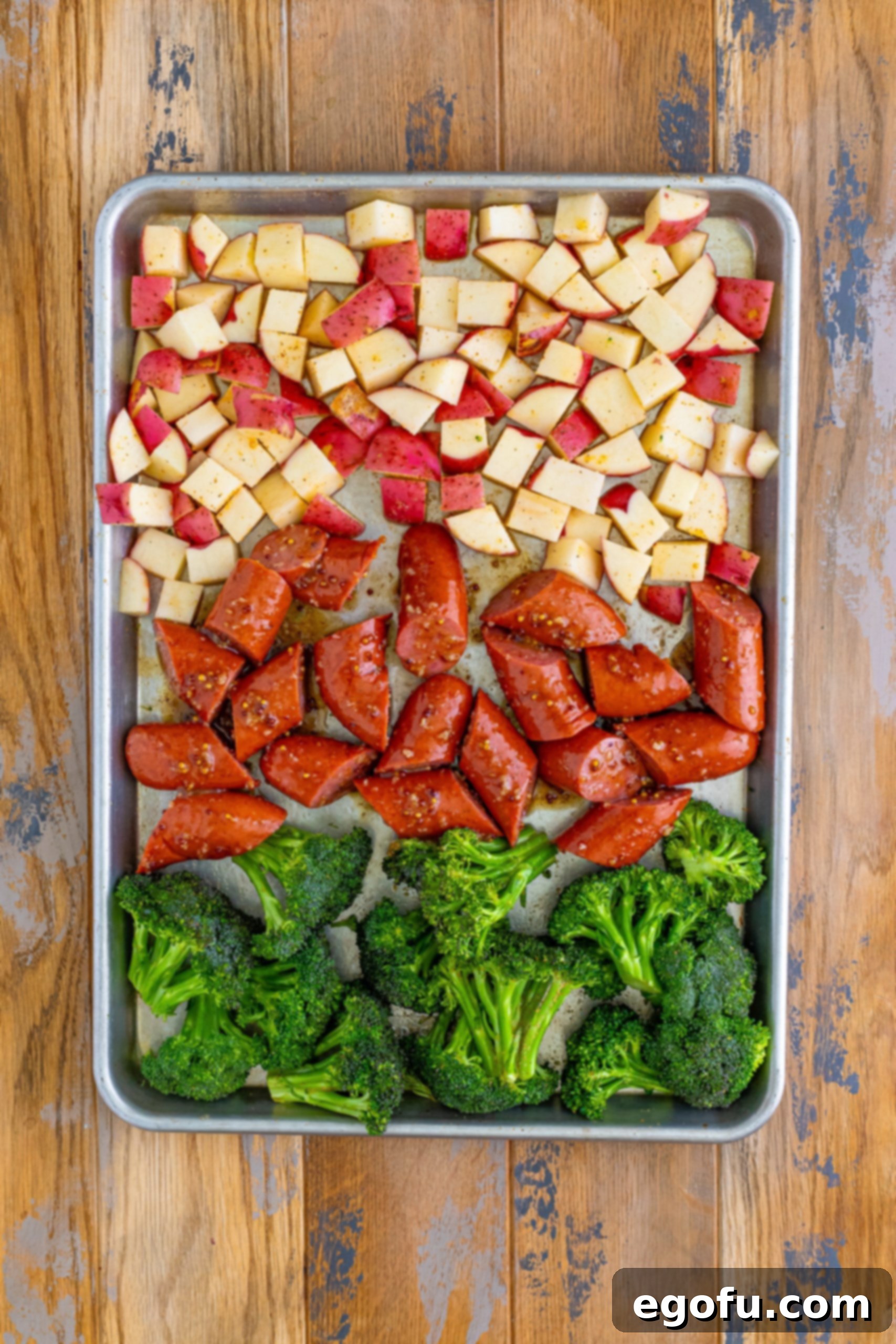Kielbasa coated with sauce, broccolini, and potatoes arranged together on a sheet pan, ready for the oven.