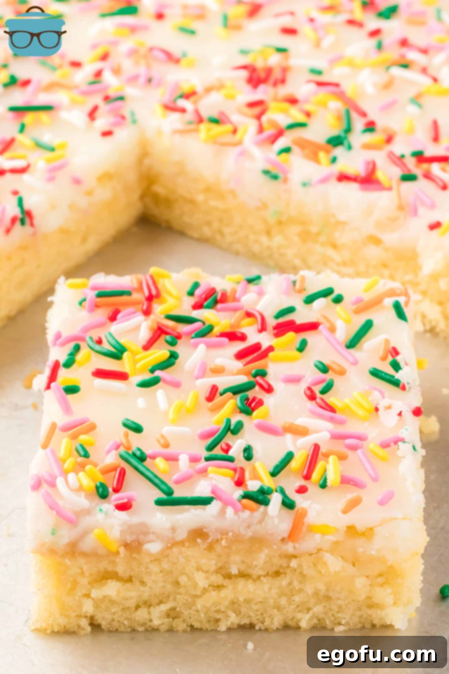 A large slice of White Texas Sheet Cake topped with sprinkles, placed on a white cake stand next to the remaining cake in its pan.