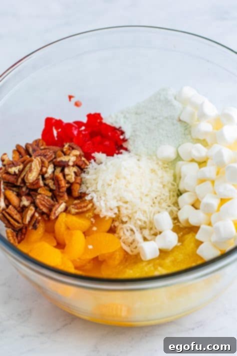 A large bowl with crushed pineapple, oranges, marshmallows, pudding mix, cherries, pecans, and coconut flakes.