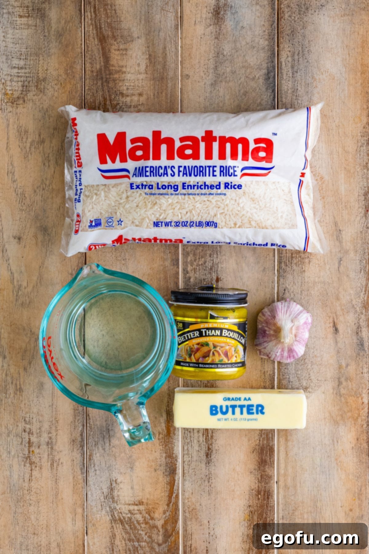 Individual ingredients for Garlic Butter Rice laid out: long-grain rice, a stick of unsalted butter, a cup of water, a jar of chicken bouillon paste, and several fresh garlic cloves.