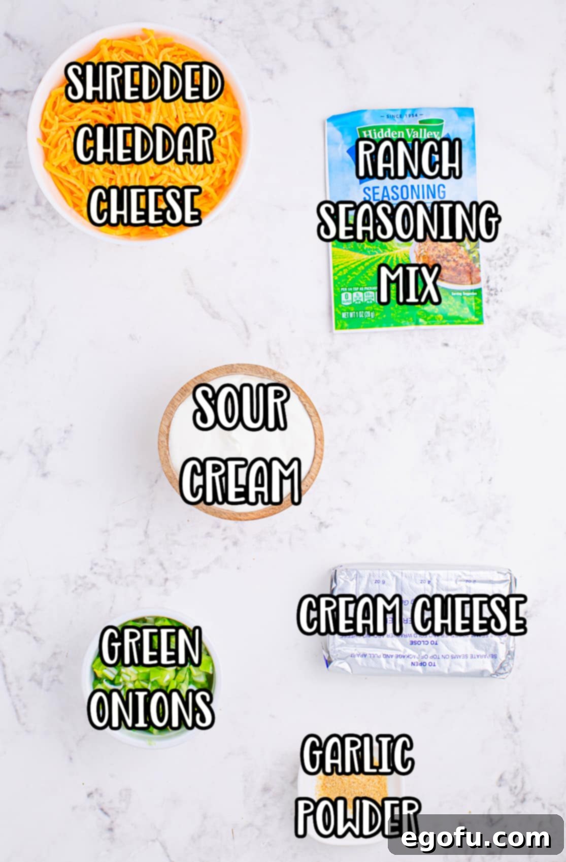 A flat lay image displaying all the key ingredients for Cheddar Ranch Dip: Ranch seasoning packet, shredded cheddar cheese, cream cheese block, sour cream container, chopped green onions, and garlic powder. 