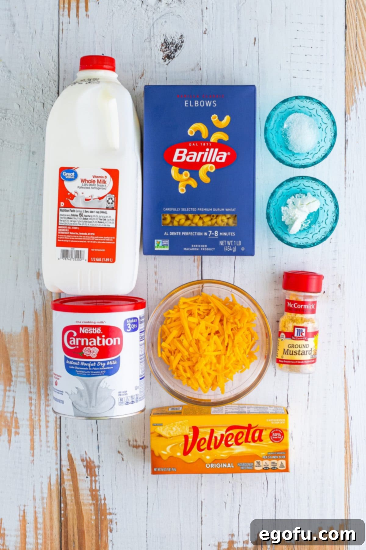 A selection of ingredients laid out: whole milk, elbow macaroni, dry ground mustard, salt, dry milk powder, Velveeta cheese, cornstarch, and sharp cheddar cheese.