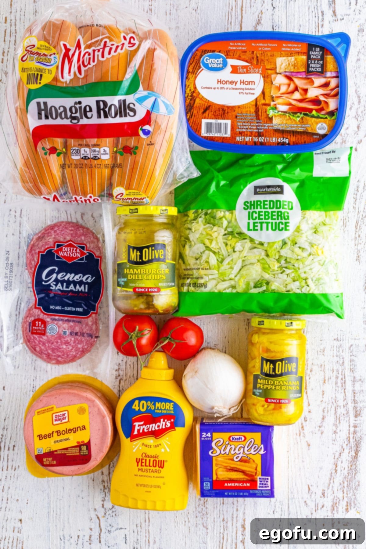 A vibrant display of fresh ingredients for Kmart Subs: soft sub rolls, bright yellow mustard, crisp shredded lettuce, ripe tomatoes, thinly sliced white onion, a trio of Genoa salami, bologna, and honey ham, creamy American cheese, tangy banana peppers, and crunchy dill pickles.