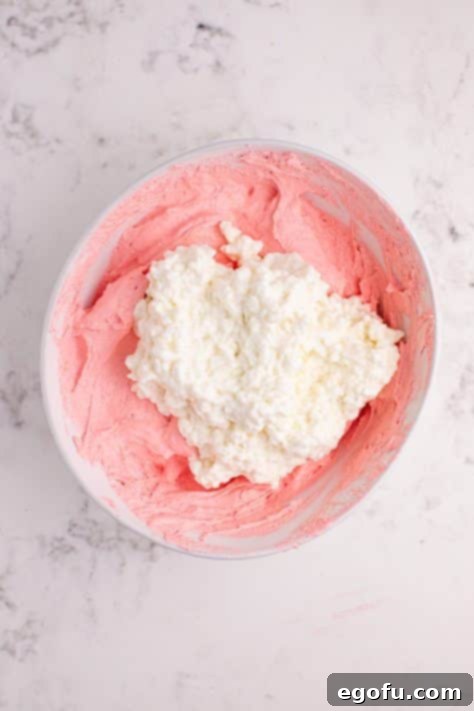 Cottage cheese being added to raspberry Jell-o, whipped topping and powdered sugar in a bowl.