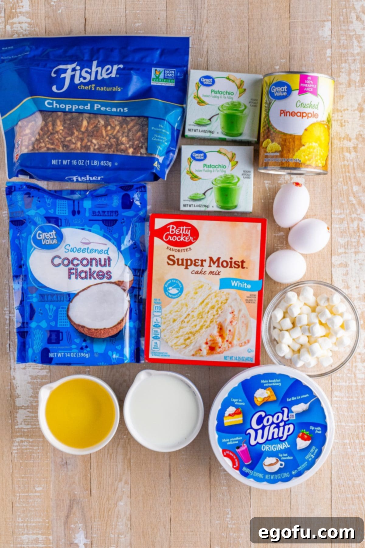 An array of Watergate Cake ingredients, including crushed pineapple, chopped pecans, white cake mix, vegetable oil, whipped topping, eggs, sweetened coconut flakes, mini marshmallows, and instant pistachio pudding mix.