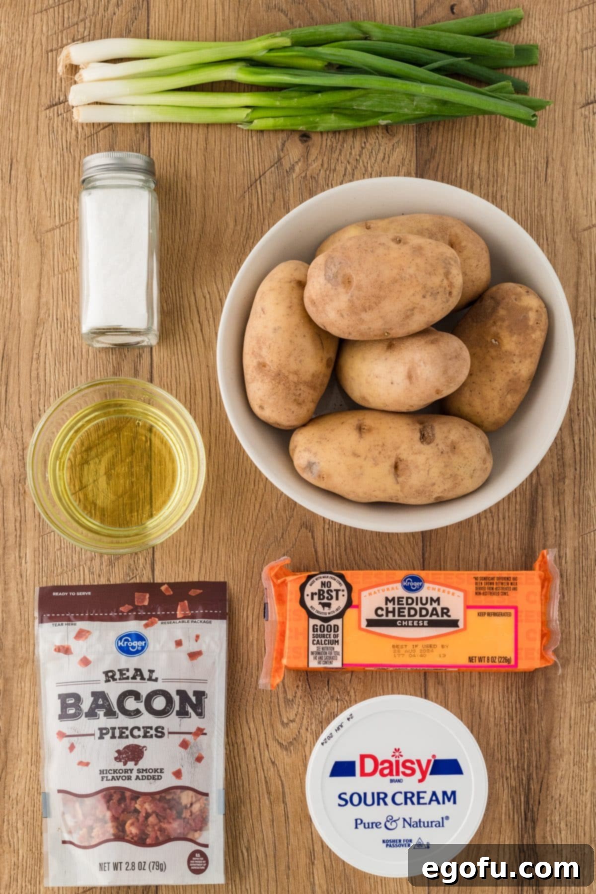 Arrangement of ingredients for Air Fryer Potato Skins: Russet potatoes, olive oil, salt, a block of cheddar cheese, bacon pieces, sour cream, and fresh green onions.