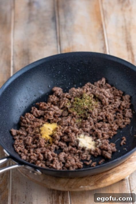 Ground beef with spices, including Italian seasoning, garlic powder, onion powder, and Parmesan cheese, mixed in a skillet.