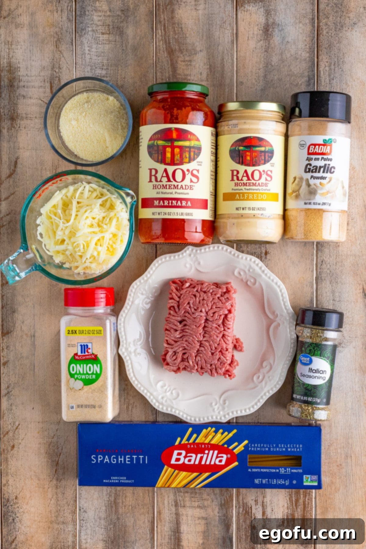 A visual display of all the ingredients needed for Baked Spaghetti for Two: Spaghetti noodles, ground beef, Italian seasoning, garlic powder, onion powder, grated parmesan cheese, jarred marinara, jarred Alfredo, and shredded mozzarella cheese arranged neatly on a surface.
