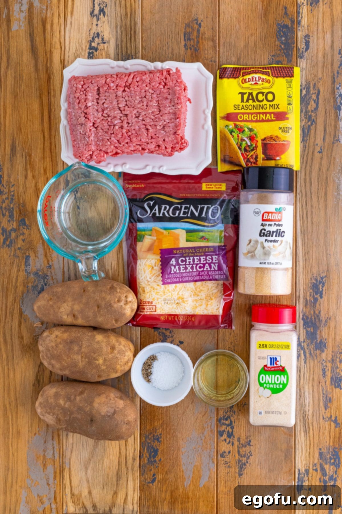 A flat lay of all the essential ingredients for Taco Potatoes: ground beef, water, taco seasoning, russet potatoes, Mexican shredded cheese, garlic powder, onion powder, salt, and pepper.