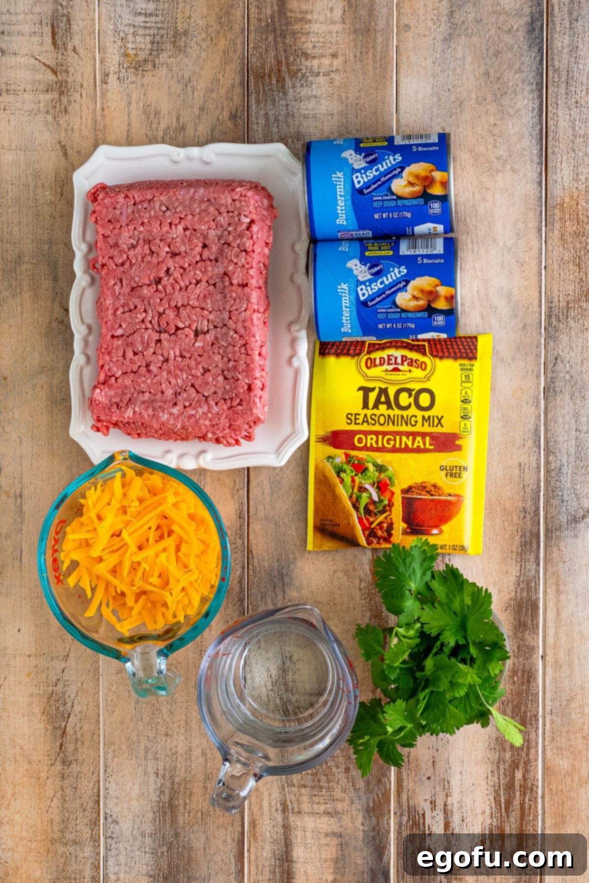 Lean ground beef, shredded cheddar cheese, fresh cilantro, taco seasoning, water, and canned biscuit dough laid out as ingredients.