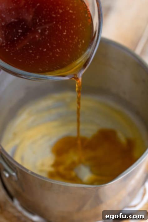 A steady stream of beef broth being poured into a saucepan with a simmering roux, actively whisked to prevent lumps and ensure a smooth gravy.