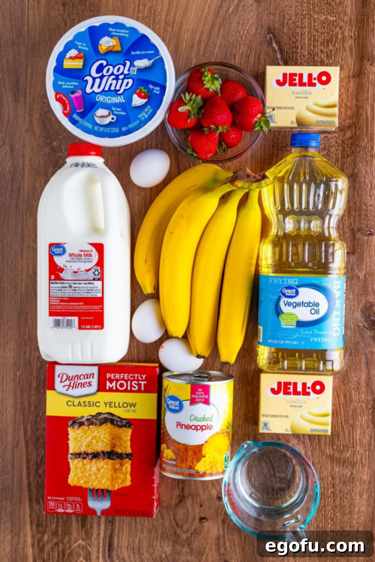 Party Perfect Bowl Cake 3 A vibrant flat lay showcasing all the essential ingredients: Cool Whip, whole milk, fresh bananas, bright red strawberries, instant vanilla pudding mix, cooking oil, yellow cake mix, eggs, and a can of crushed pineapple, neatly arranged on a white surface.