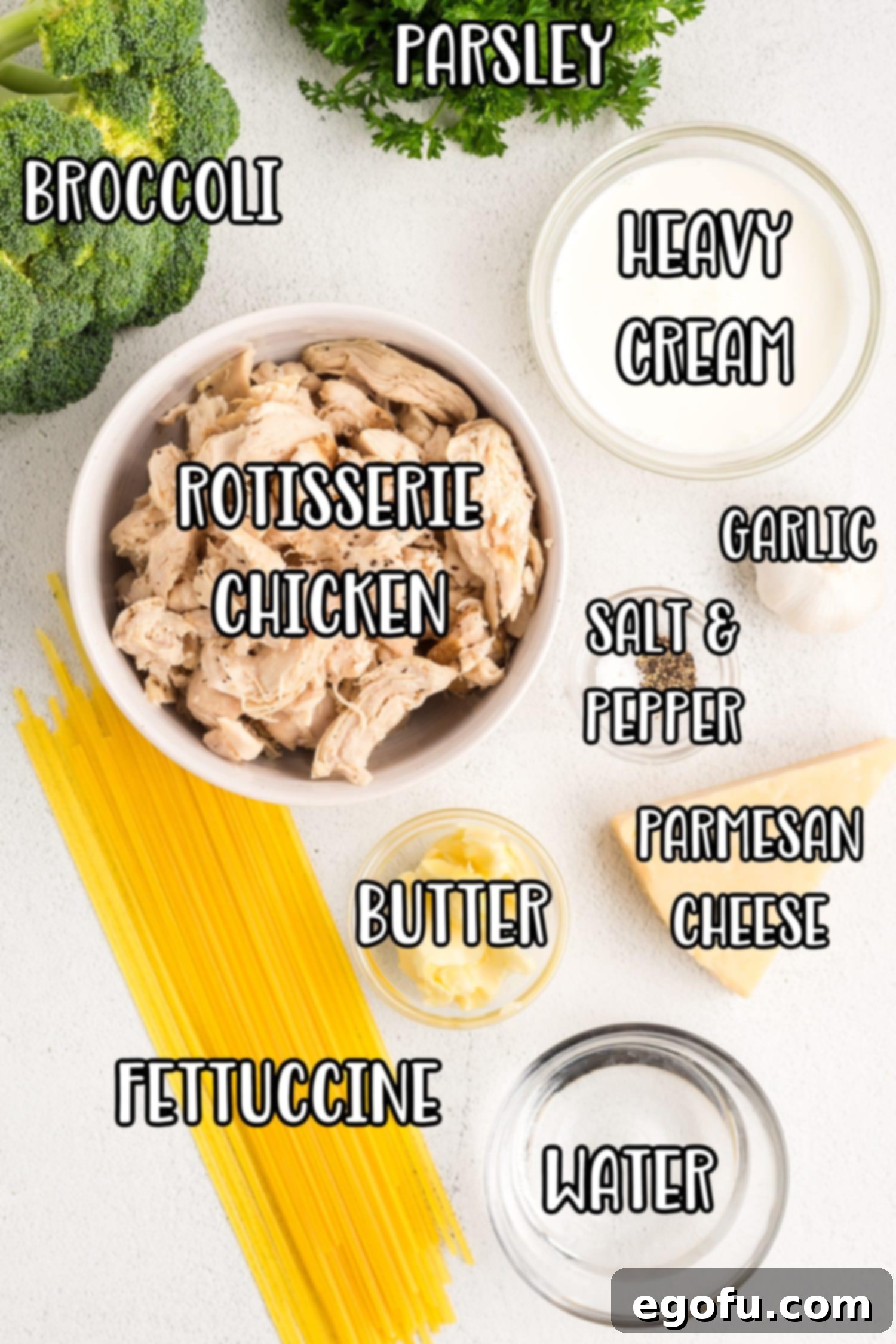 An array of ingredients for Chicken Broccoli Alfredo: rotisserie chicken, broccoli, fettuccine, water, black pepper, salt, unsalted butter, garlic, parmesan cheese, heavy cream, and chopped parsley arranged on a rustic surface.