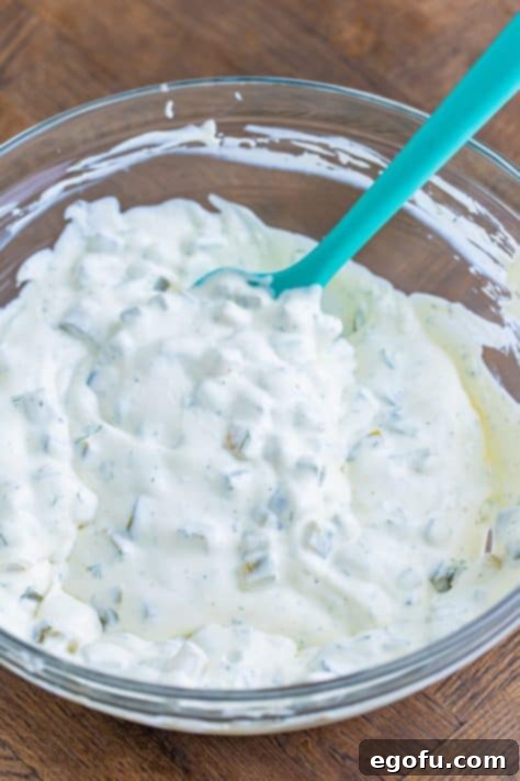 A bowl of creamy pickle dip covered with plastic wrap, chilling in the fridge.