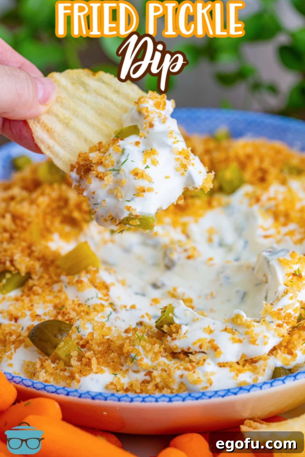 A hand holds a chip generously coated with Fried Pickle Dip, ready to be enjoyed, showing the rich texture and topping.