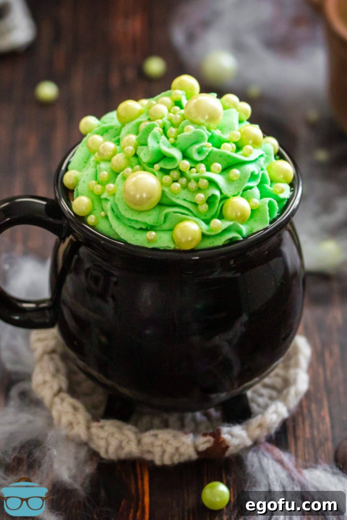 Bewitching Brew Hot Chocolate 2 A cauldron-style mug filled with dark Witches Brew Hot Chocolate, topped with fluffy green whipped cream and whimsical green sphere sprinkles, ready for a Halloween celebration.