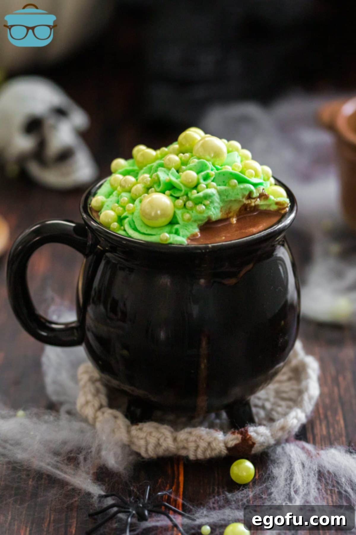 Bewitching Brew Hot Chocolate 7 A black cauldron mug with rich, dark Witches Brew Hot Chocolate, topped generously with bright green whipped cream and whimsical green sphere sprinkles, perfect for Halloween.