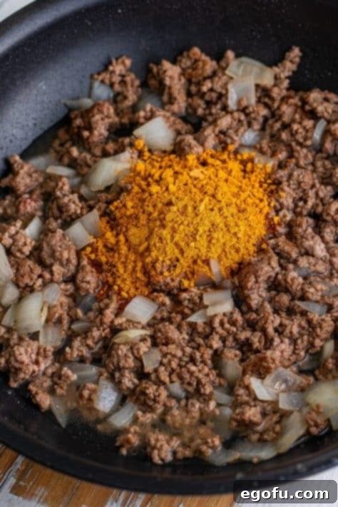 Taco seasoning being sprinkled over the cooked ground beef and onions in a skillet, ready to be stirred with water to form the flavorful filling.