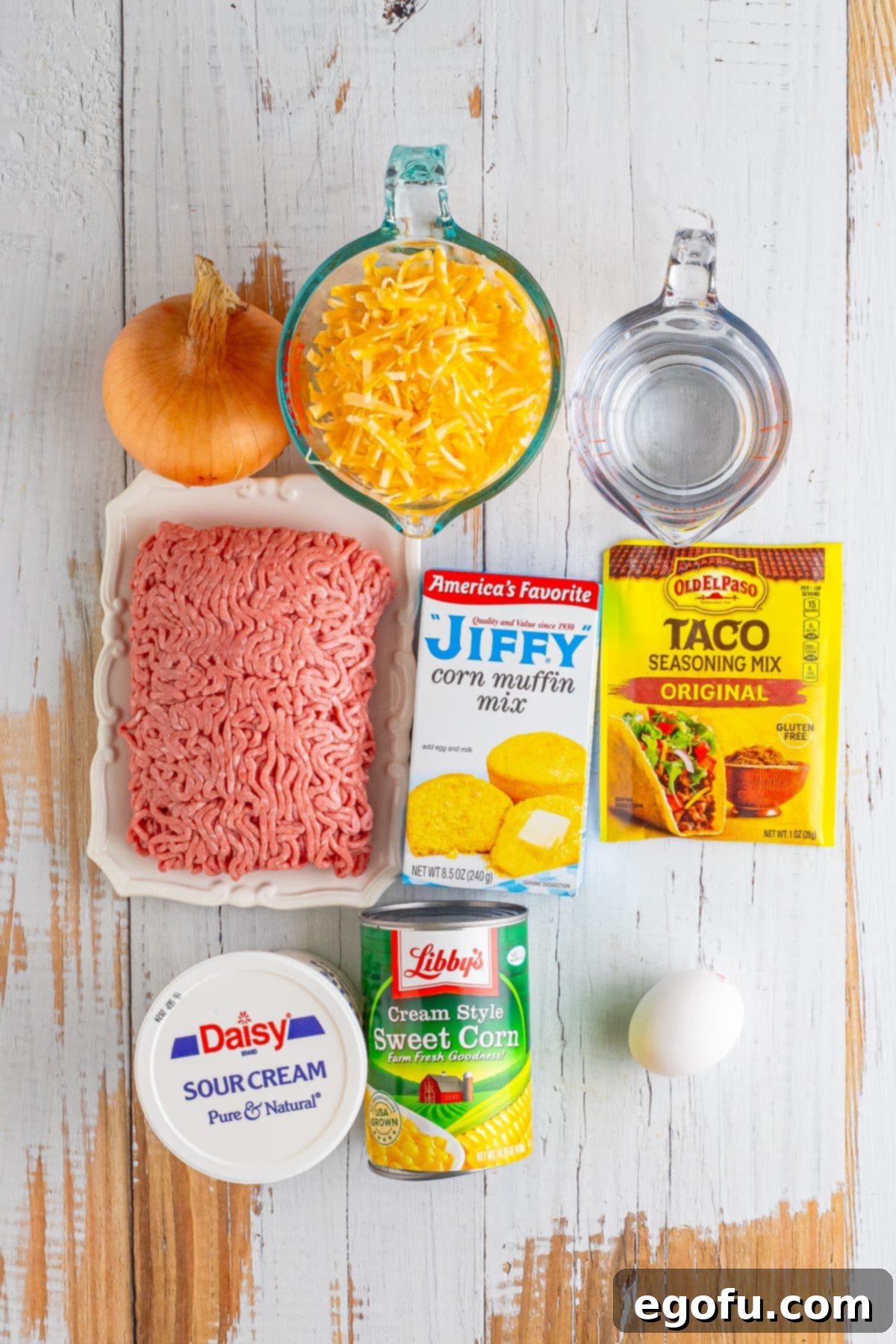 A flat lay photograph displaying all the fresh and packaged ingredients needed for Tamale Pie: ground beef, creamed corn, cornbread mix, sour cream, egg, onion, taco seasoning, water, and Colby Jack cheese.