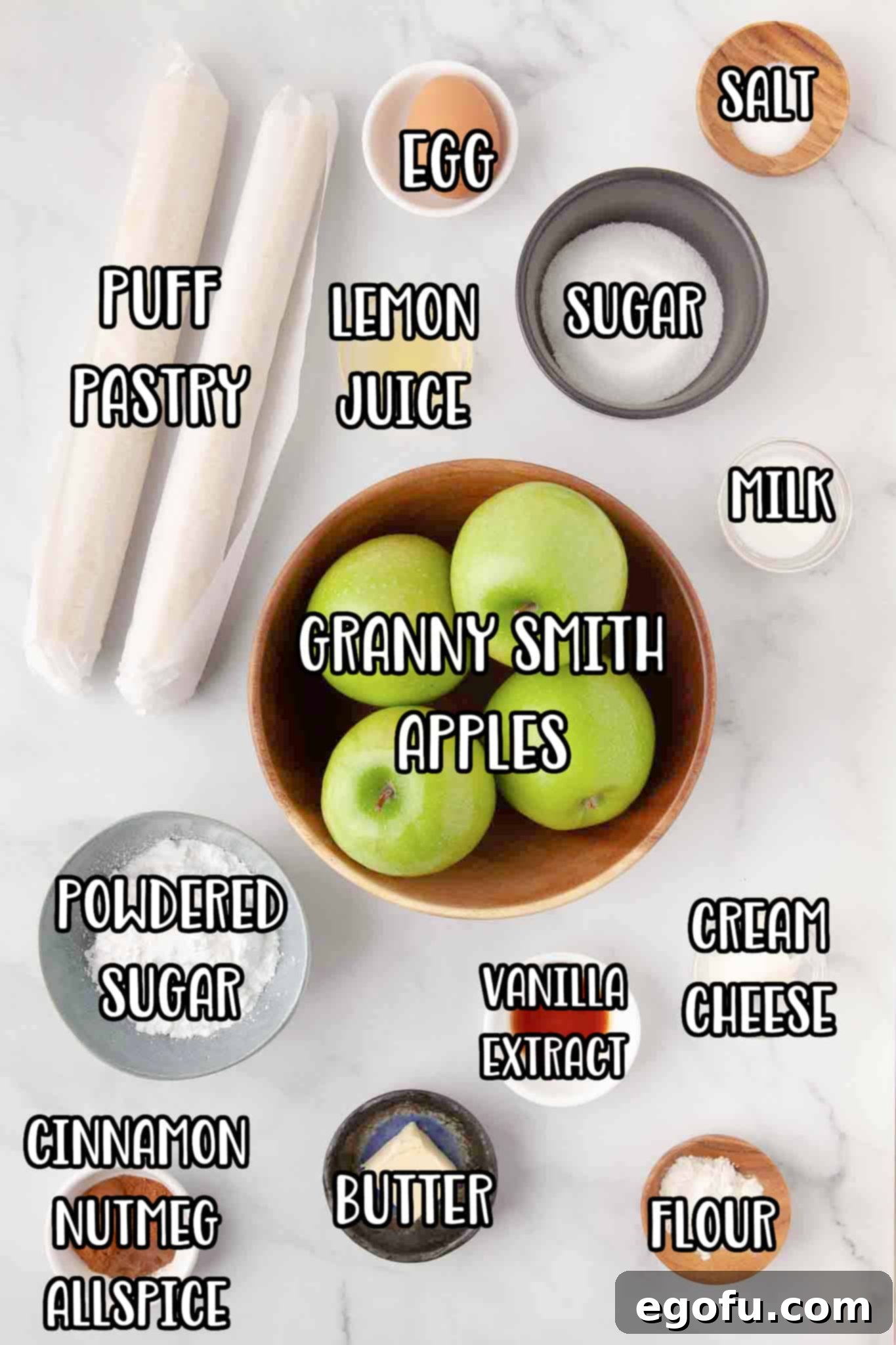 Salt, egg, puff pastry, lemon juice, milk, Granny Smith apples, cream cheese, vanilla extract, powdered sugar, butter, flour, cinnamon, nutmeg, and all spice arranged on a surface.