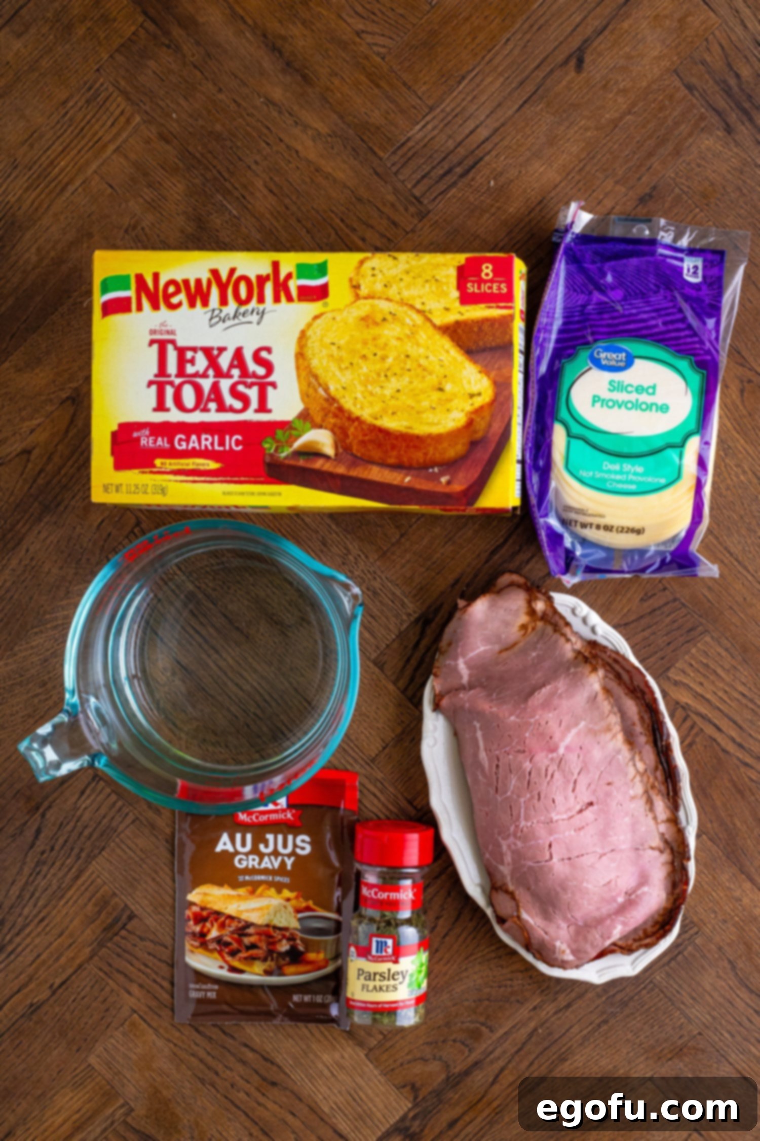 All the essential ingredients for making Texas Toast French Dips laid out neatly: frozen garlic Texas Toast, a packet of au jus gravy mix, a measuring cup with cold water, thinly sliced deli roast beef, provolone cheese slices, and a small dish of dried parsley.