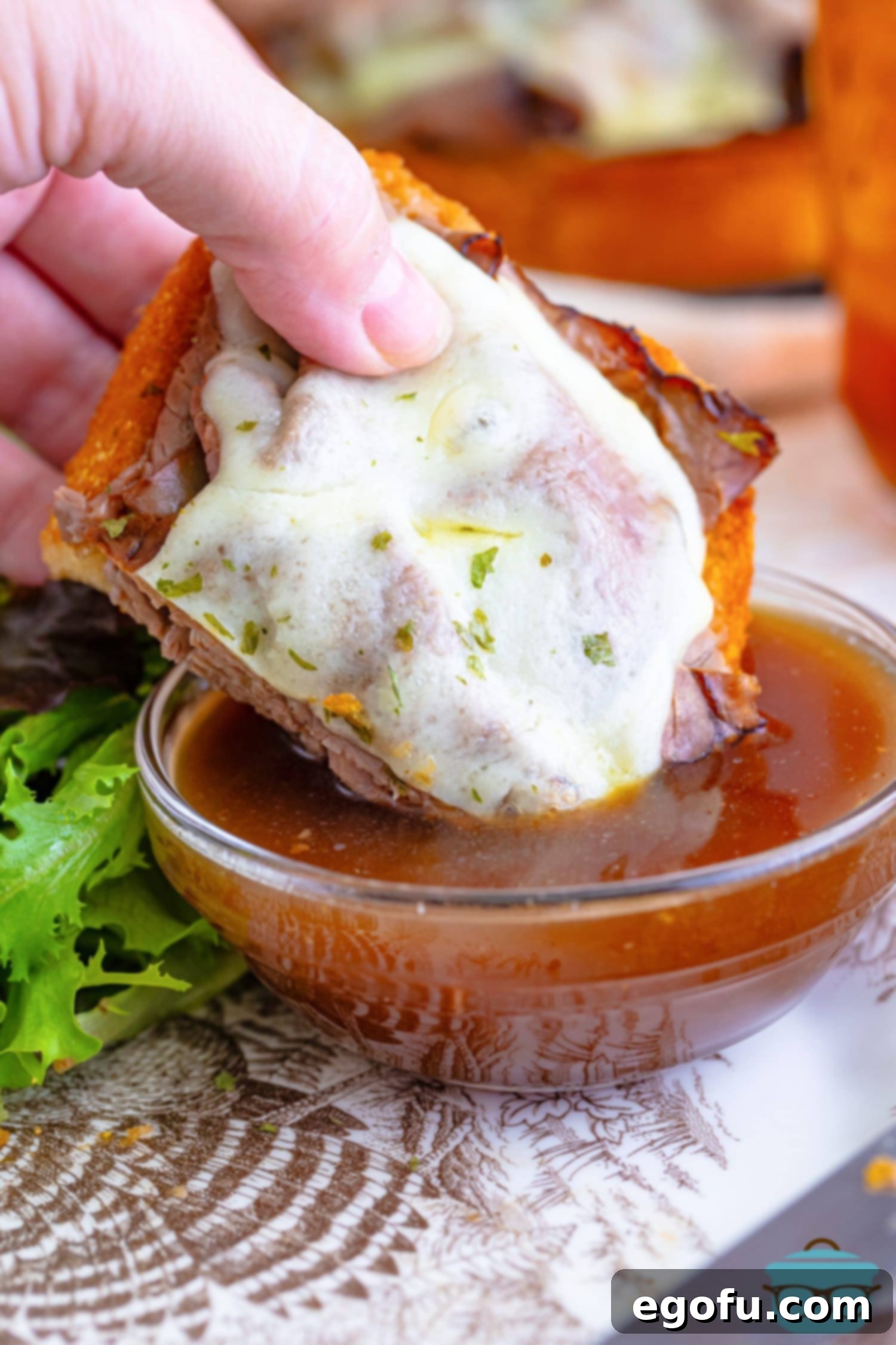 A close-up shot of a hand dipping a freshly prepared Texas Toast French Dip into a small, steaming bowl of rich, brown au jus gravy, highlighting the interactive and flavorful dining experience.