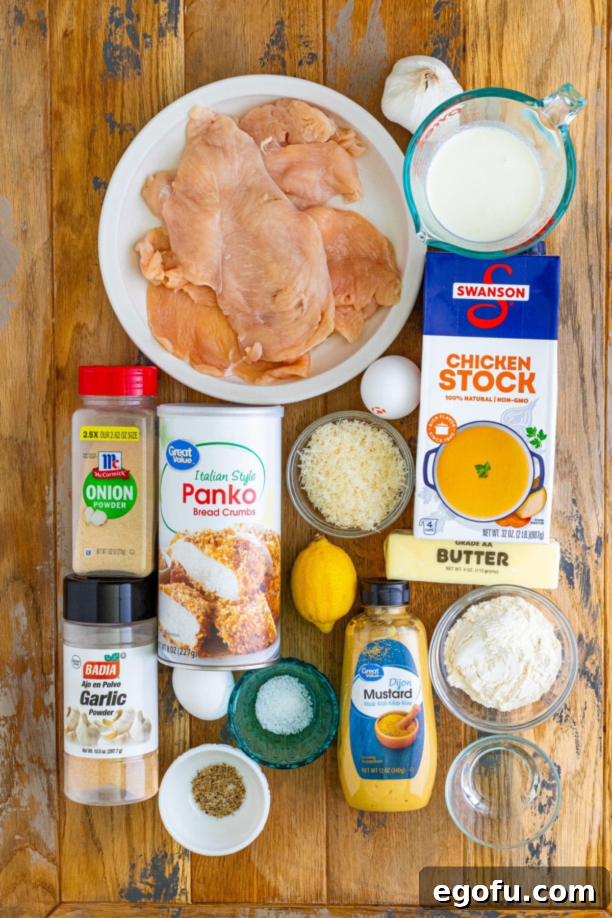 An organized array of fresh ingredients including all-purpose flour, garlic powder, onion powder, salt, pepper, eggs, water, Italian Panko breadcrumbs, fresh lemon, grated Parmesan cheese, raw thin-cut chicken breasts, butter, fresh garlic cloves, heavy cream, and Dijon mustard, artfully laid out on a clean surface.