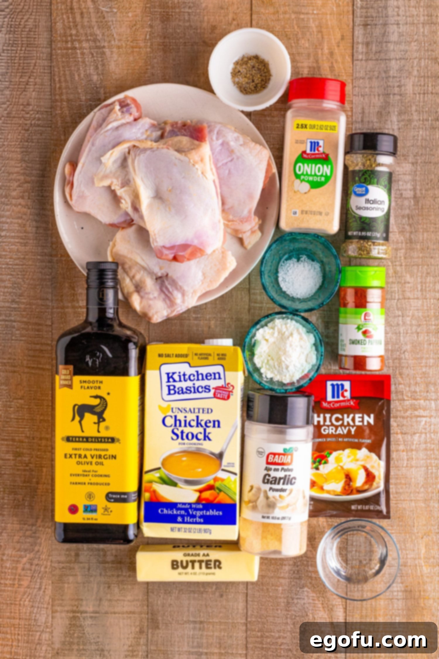 A flat lay of all the ingredients needed: chicken thighs, individual spices (salt, garlic powder, onion powder, paprika, Italian seasoning, pepper), olive oil, chicken stock, chicken gravy mix, butter slices, cornstarch, and water.
