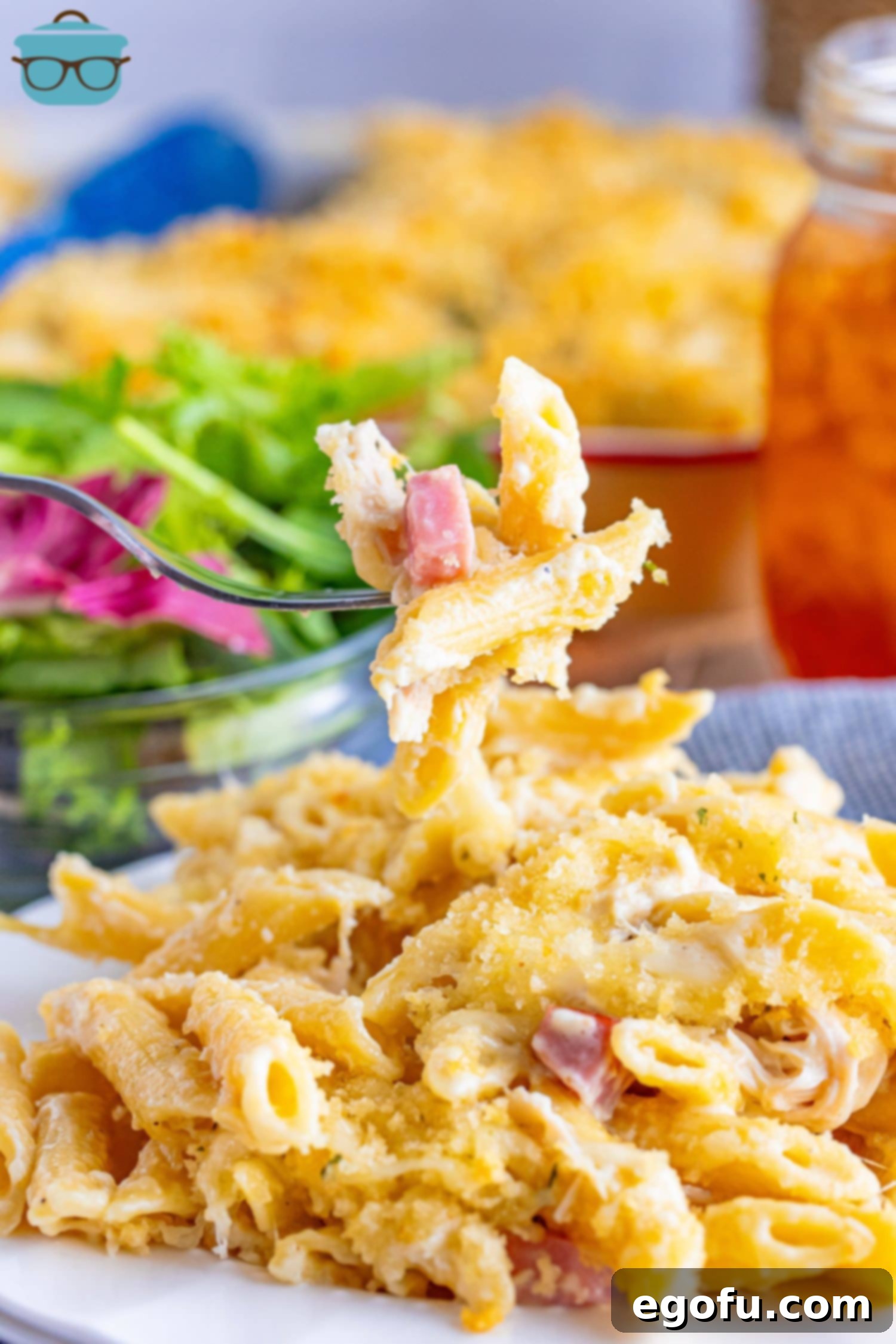 A fork holding a delicious bite of Malibu Chicken Casserole, showing the layers of pasta, chicken, ham, and melted cheese with a crispy topping.