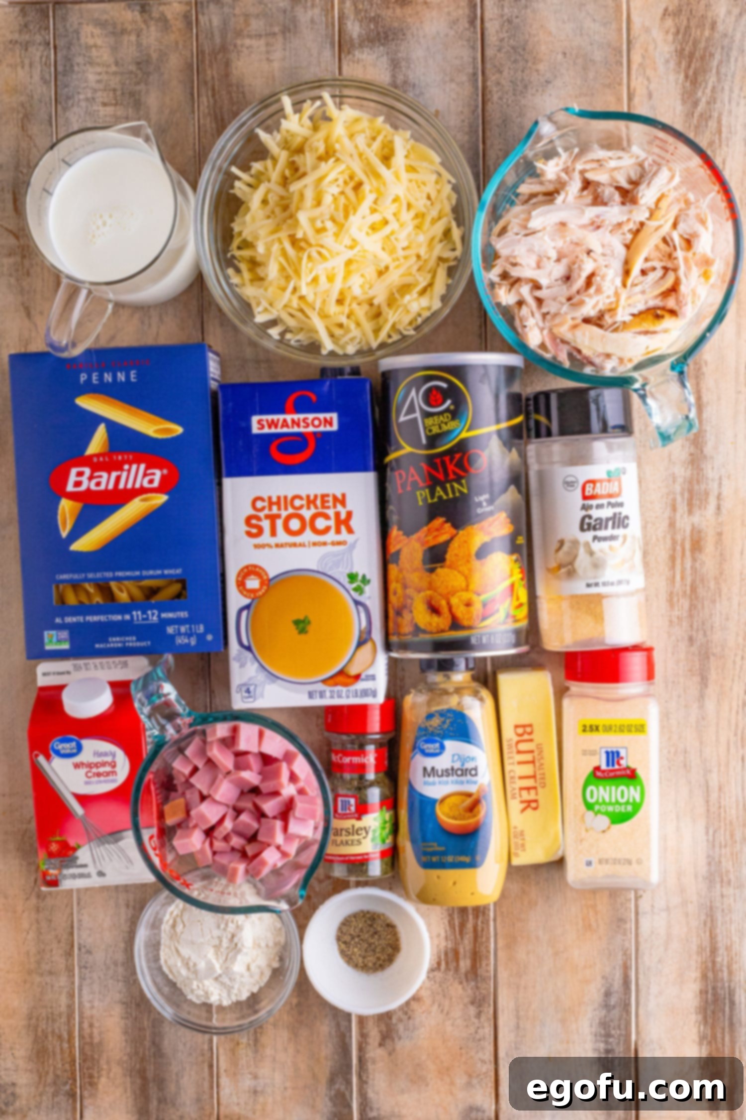An array of ingredients for Malibu Chicken Casserole laid out: shredded chicken, Swiss cheese, diced ham, chicken stock, butter, flour, garlic powder, onion powder, pepper, whole milk, heavy cream, Dijon mustard, dried parsley, and panko bread crumbs.