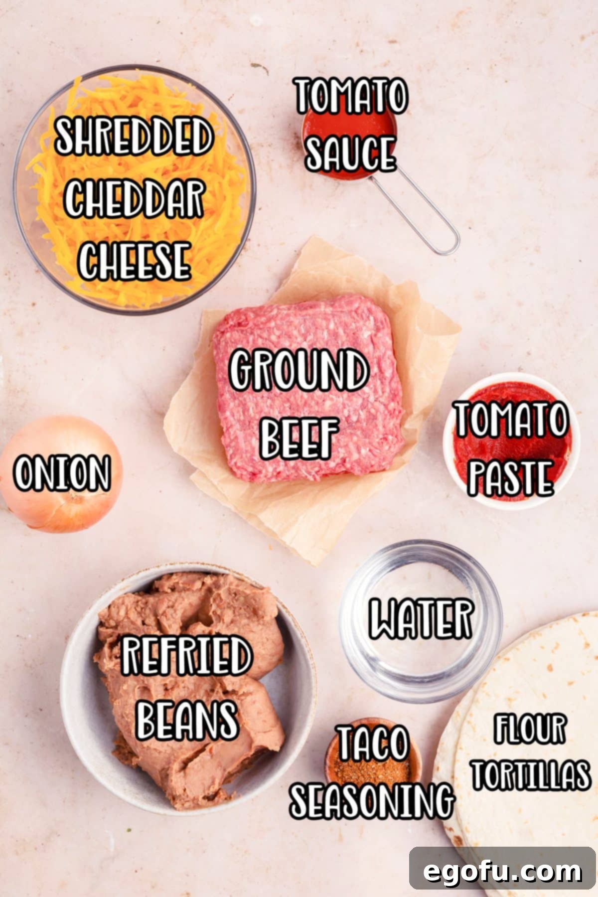 A vibrant flat lay of all the essential ingredients: shredded cheddar cheese, a packet of taco seasoning, raw ground beef, finely diced onion, tomato paste, canned refried beans, water, and stacks of soft flour tortillas.